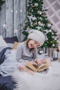 young girl with knit hat in front of christmas tree laughing with book